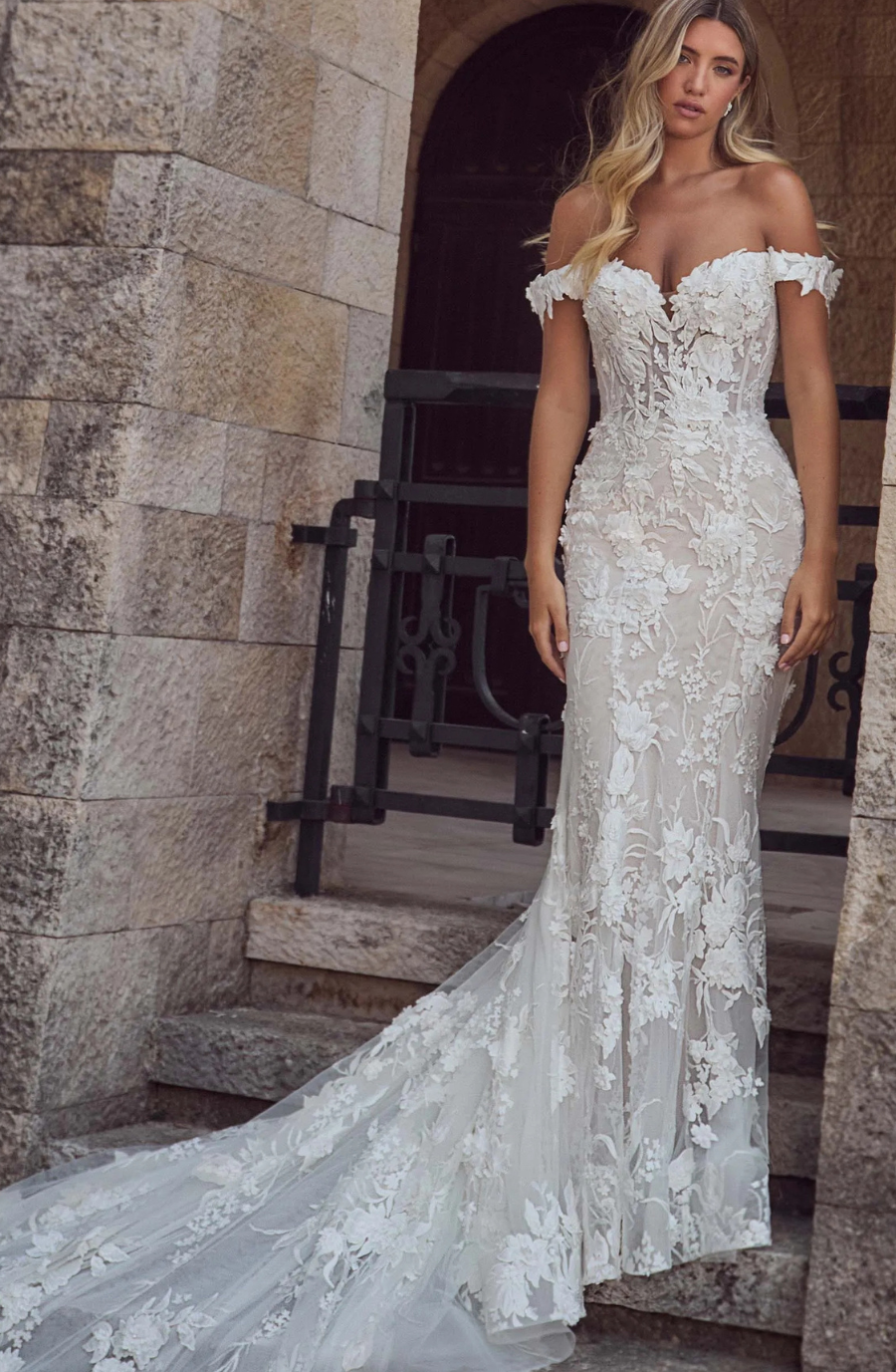 A woman wearing an elegant off-the-shoulder wedding dress with floral lace details, standing on stone steps in a historic setting.
