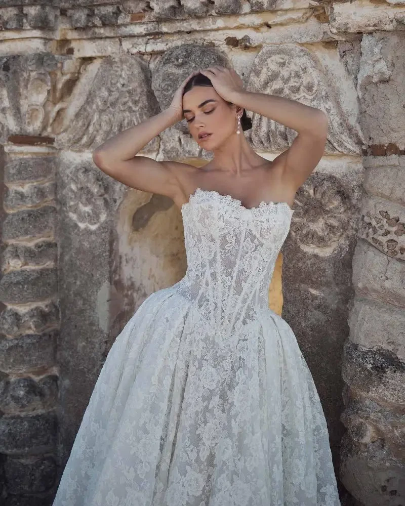 A woman in a lacy, strapless white wedding gown stands with eyes closed, hands on her head, against an ornate, weathered stone wall.