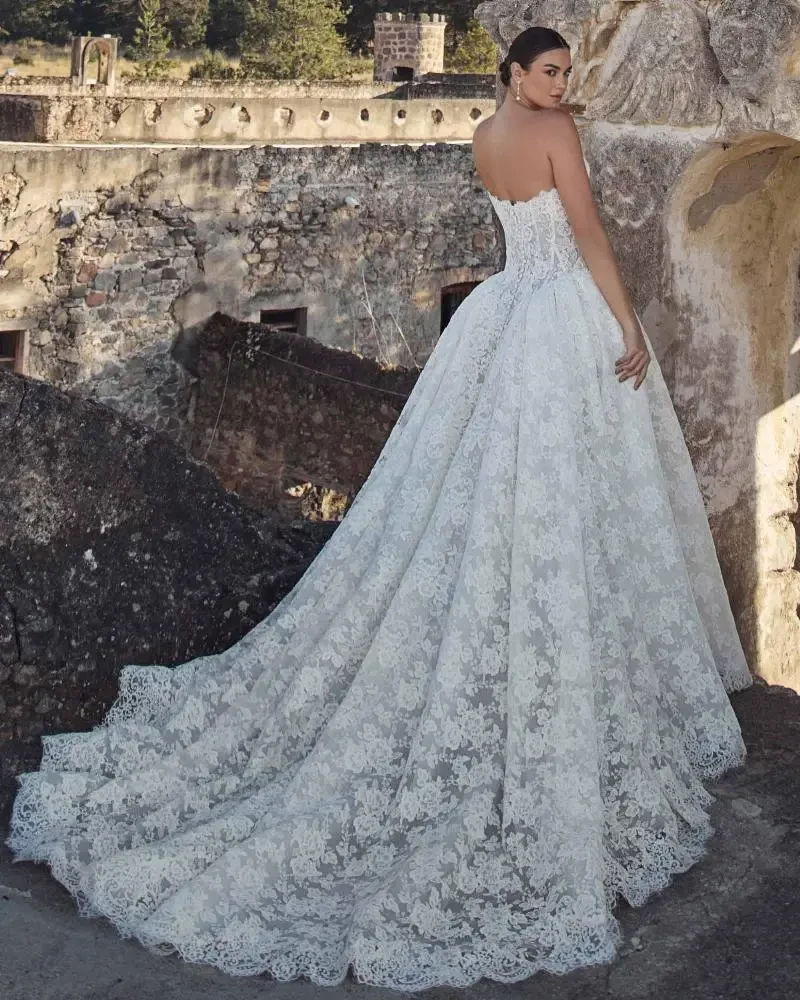 A woman in a strapless, lace wedding gown poses against a background of weathered stone ruins. The dress features a long, ornate train, adding elegance.