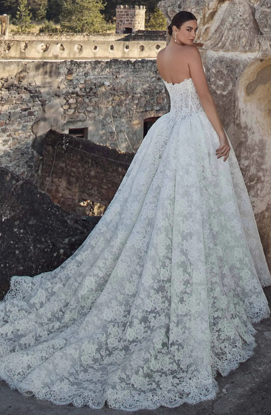 A woman in a strapless white wedding dress with a long, flowing lace train, standing with her back to the camera against an old stone wall.