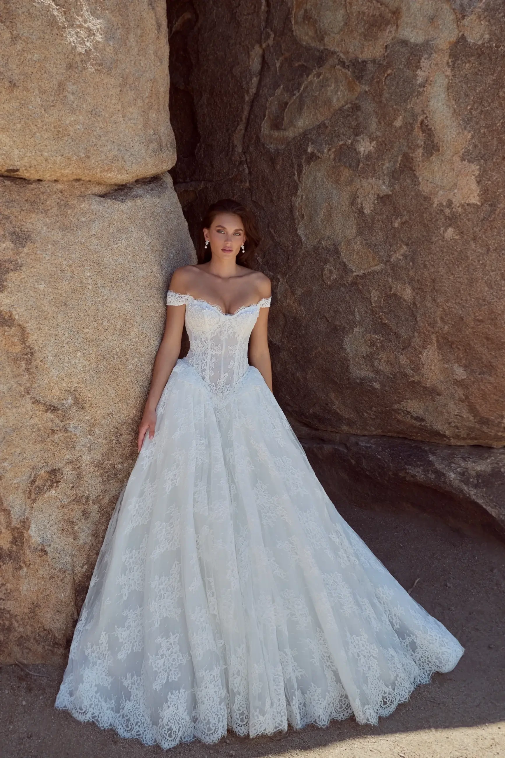 A woman in an off-the-shoulder white wedding dress stands near a large rock formation. The dress features intricate lace details and a full skirt.
