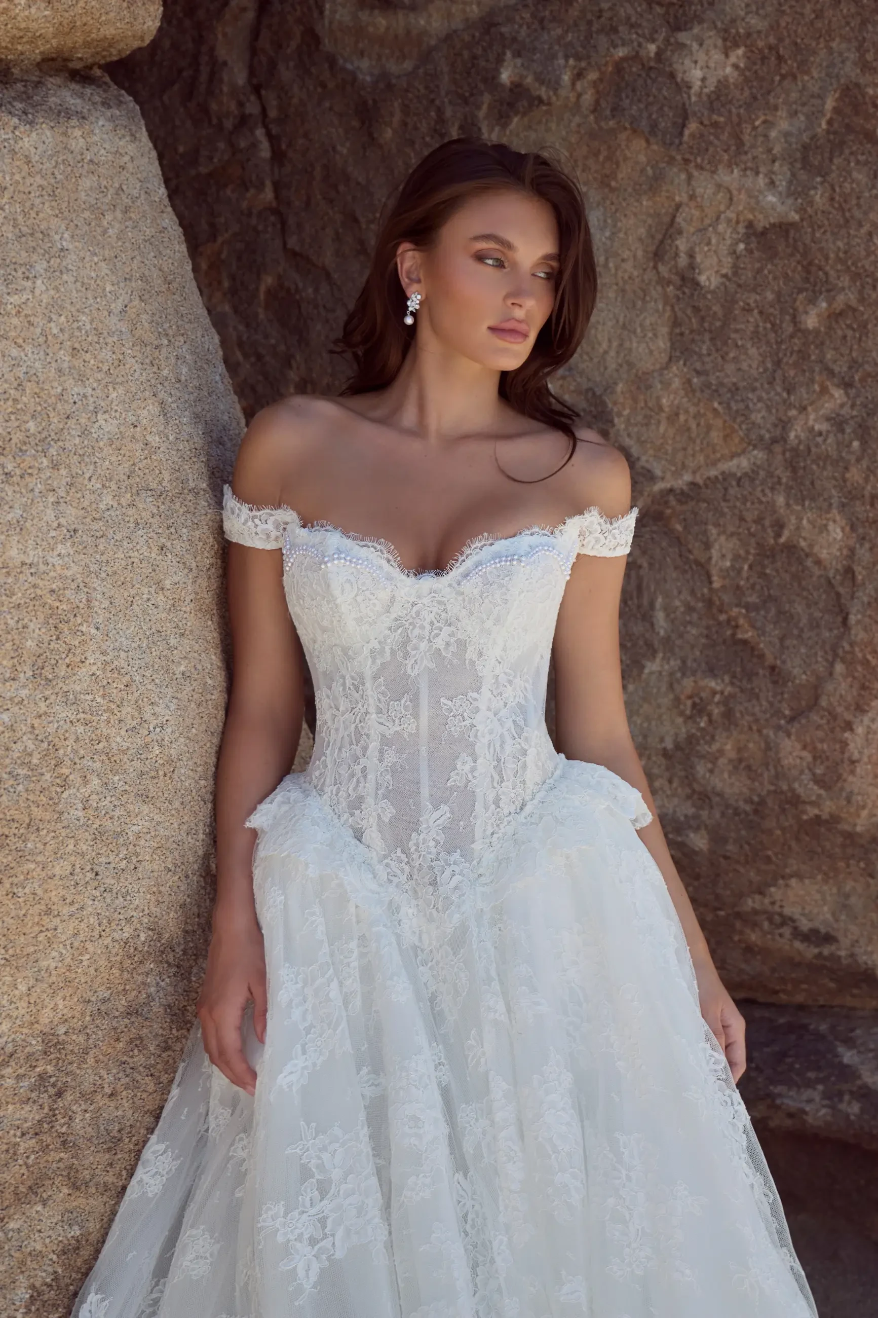 A woman in an elegant off-shoulder wedding gown with intricate lace detailing, standing against a rocky backdrop.