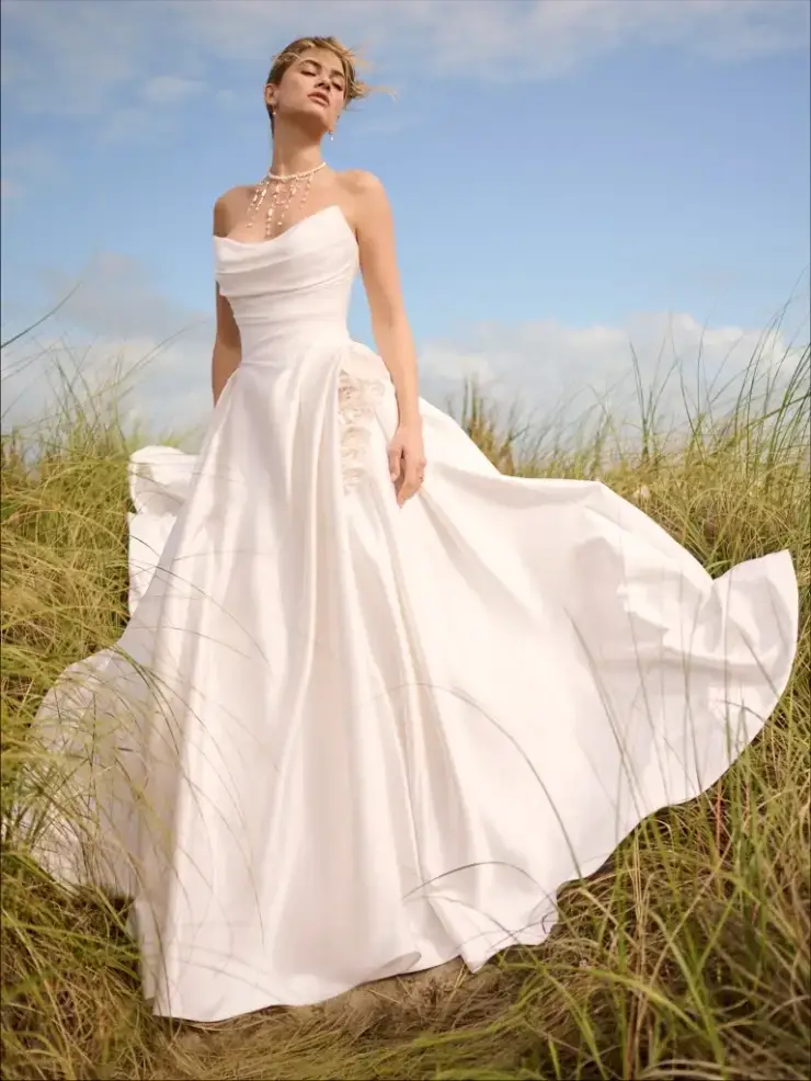 A woman wearing a flowing white dress stands in a field of tall grass, with a blue sky and wispy clouds in the background.