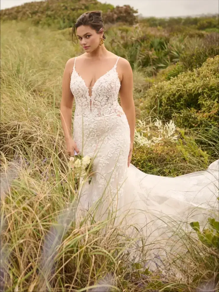 A woman in a bridal gown stands confidently in a grassy area, holding a bouquet of flowers. The dress features intricate detailing and has a flowing train, while she portrays an elegant and serene expression.