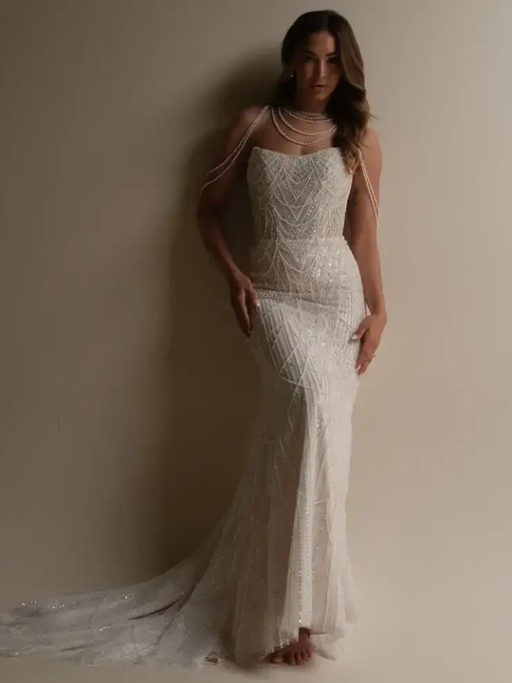 A woman wearing a stunning white dress with intricate beadwork and embellishments, posing against a neutral background. The dress has a fitted silhouette and a flowing train, complemented by layered pearl necklaces.