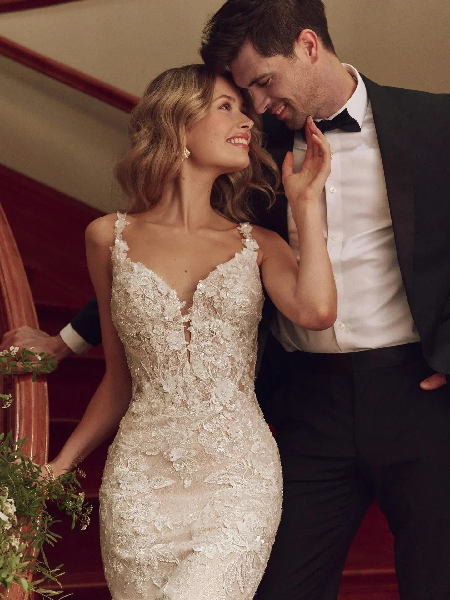 A bride in a lace wedding gown and a groom in a black suit share a joyful, intimate moment on a staircase. The scene is warm and romantic.