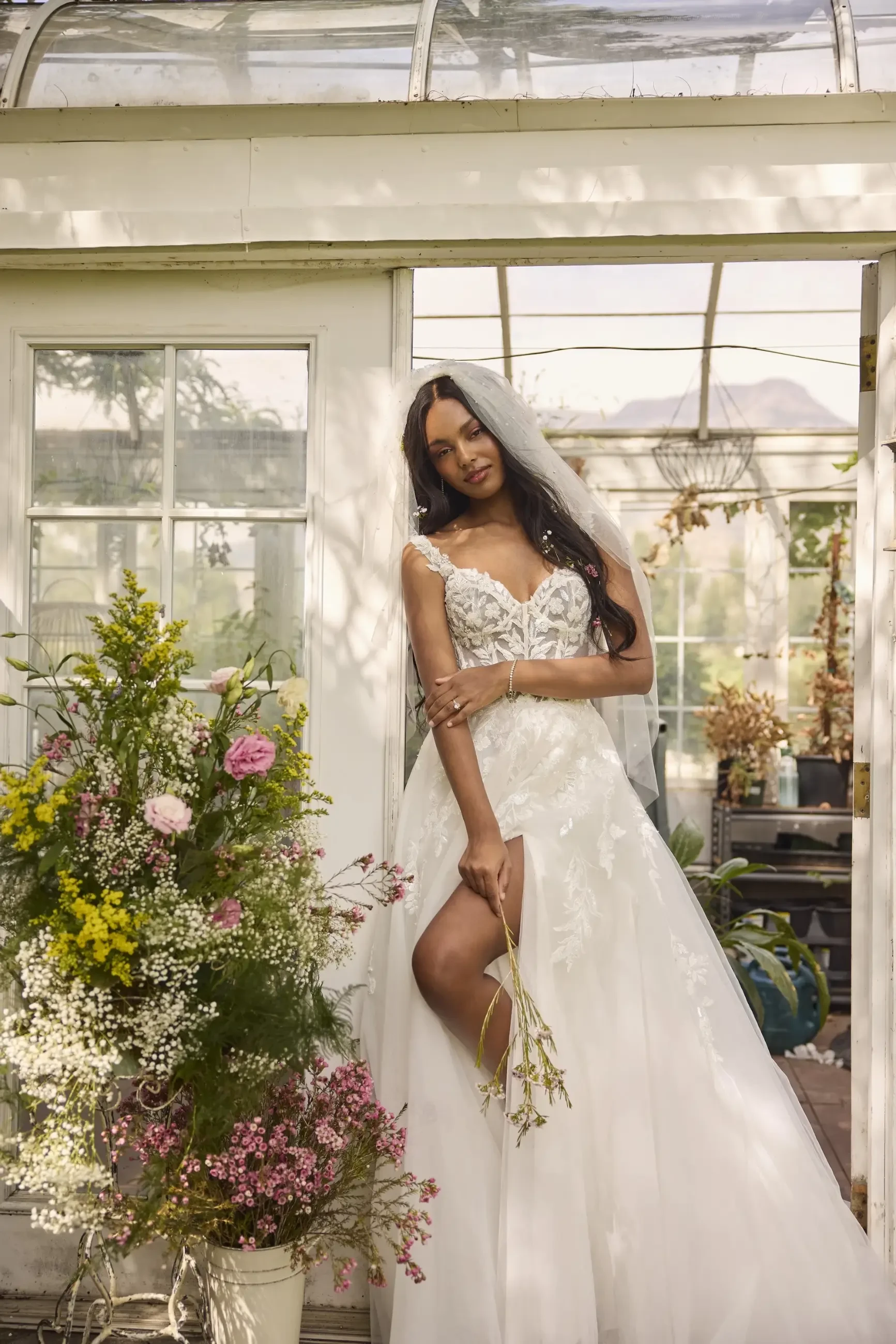 Bride in a lace wedding dress and veil stands by a flower-filled greenhouse. She holds a bouquet, exuding elegance and joy in a sunlit garden setting.