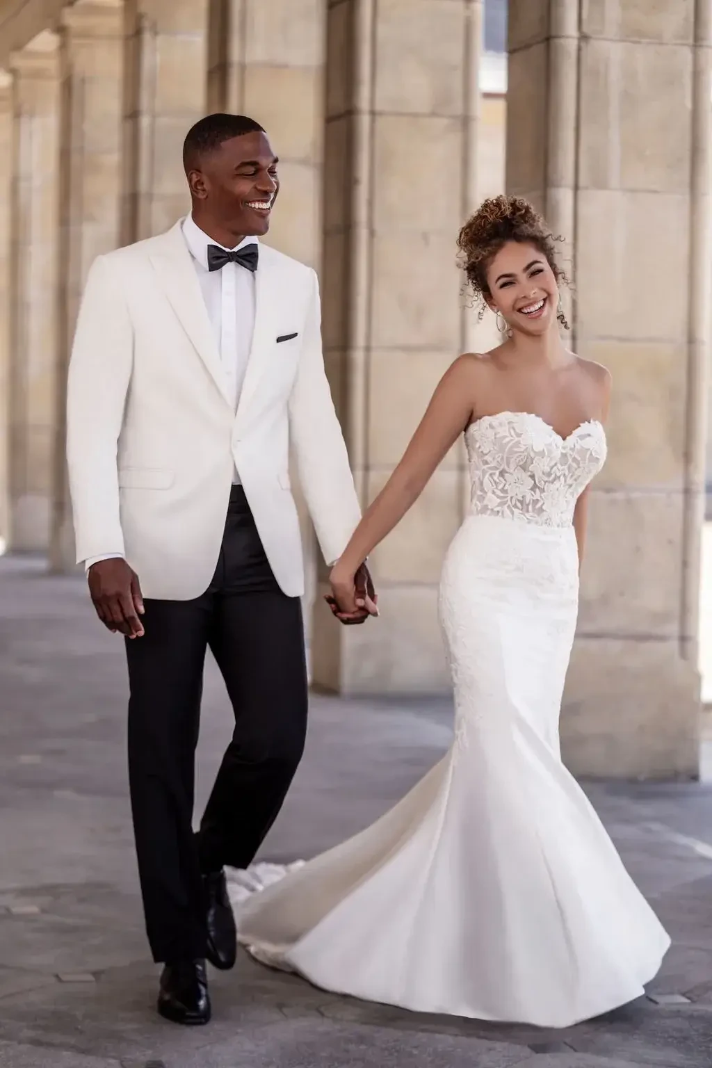 A joyful couple walks hand in hand. The groom wears a white tuxedo and black pants, while the bride dons a fitted lace wedding gown, smiling brightly.