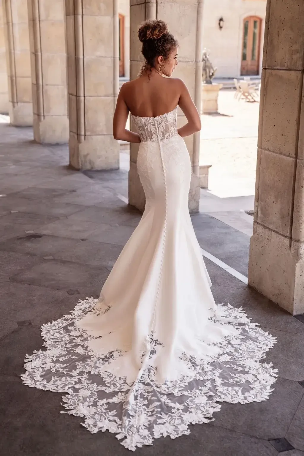 Bride in elegant strapless wedding gown with lace train stands in a stone corridor. The atmosphere is serene and timeless.