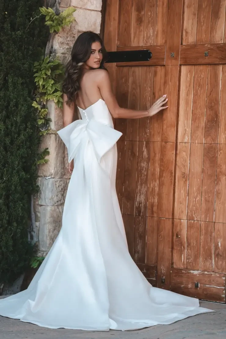 A woman in an elegant white wedding dress with a large bow stands by a wooden door. She gazes back, surrounded by greenery, exuding a serene elegance.