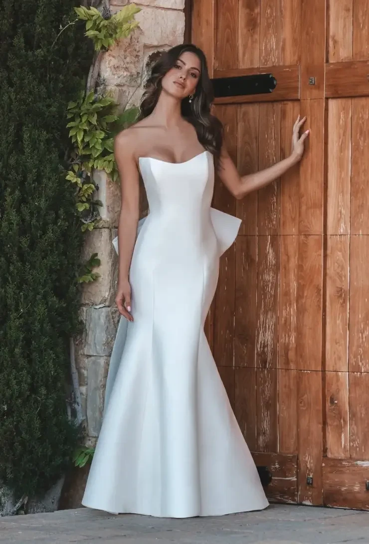 A woman in an elegant, strapless white gown poses confidently by a wooden door, surrounded by stone and green foliage, exuding grace and sophistication.
