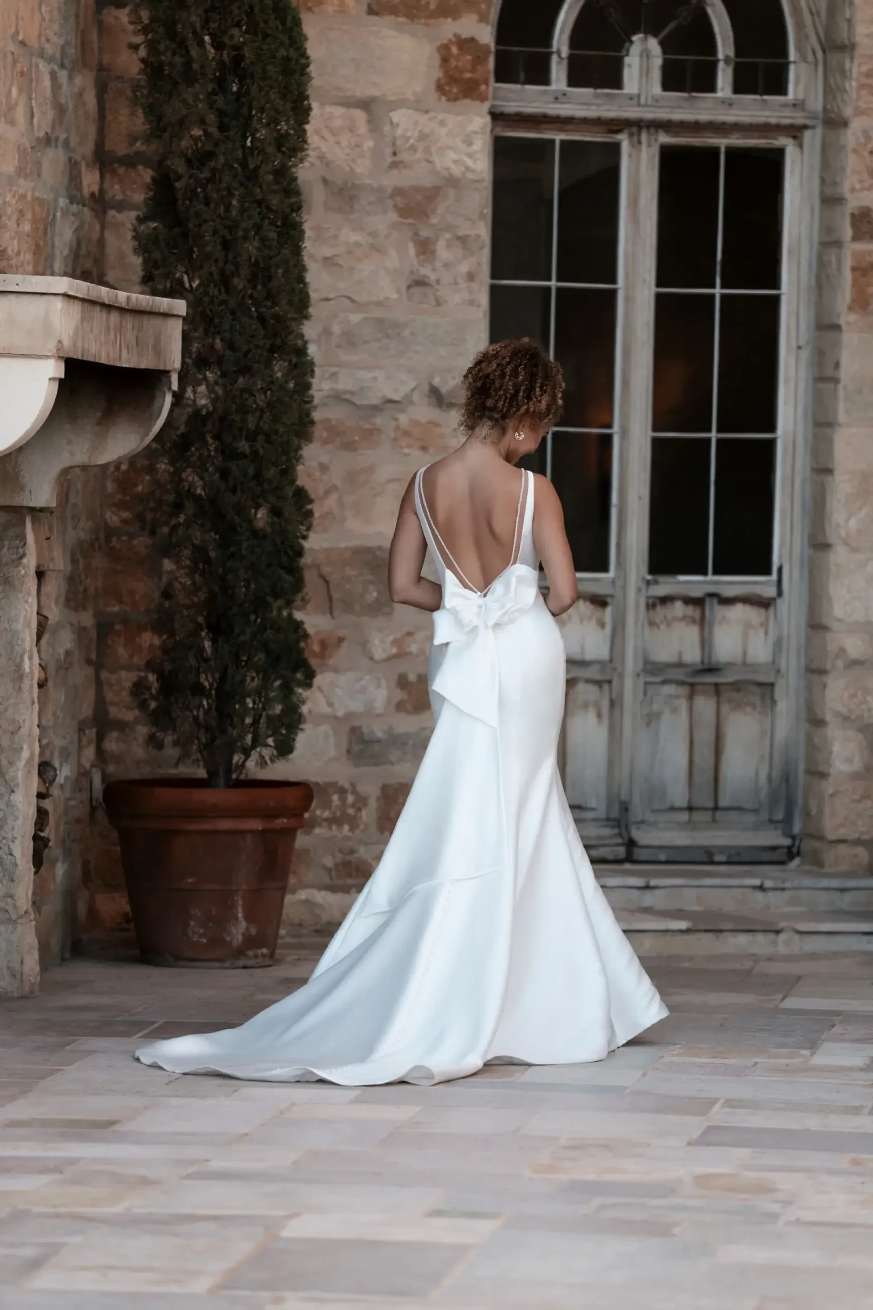 A woman in a white backless gown stands facing an old stone wall with a rustic door. A potted plant adds elegance to the serene, timeless setting.