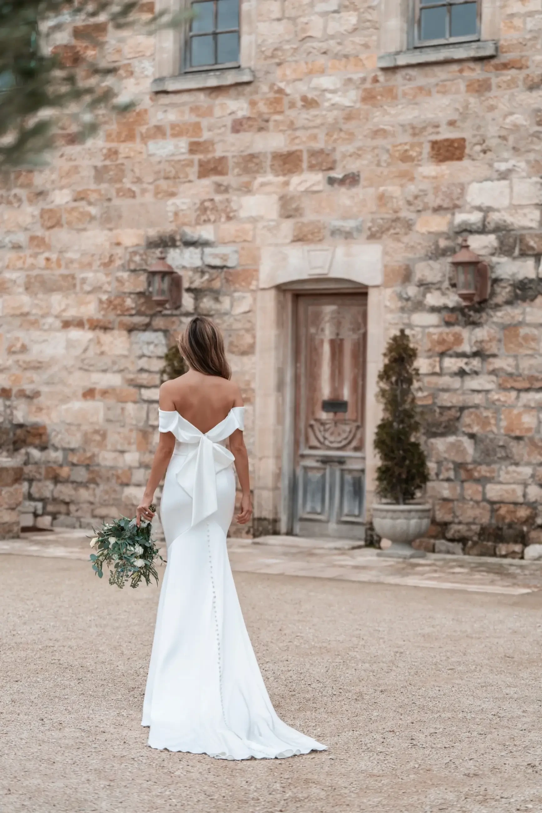 Model is wearing a chic satin sheath wedding dress with off-the-shoulder straps and a statement bow accent at the back.