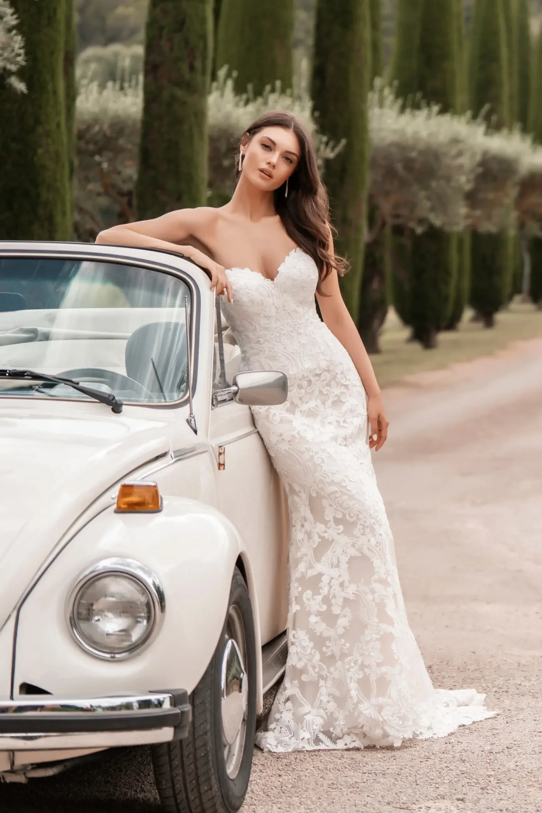 A woman in an elegant, strapless white lace wedding gown leans against a vintage white car. Behind her are tall cypress trees lining a serene path. The scene conveys romance and sophistication.