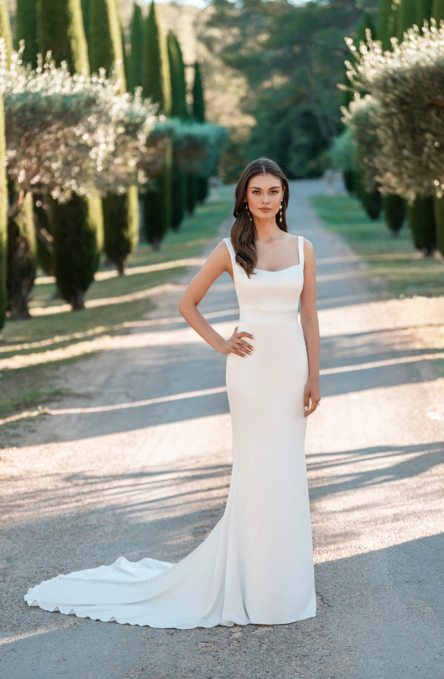A woman in a sleek white gown stands on a tree-lined path, exuding elegance and serenity. The sunlight casts soft shadows around her.