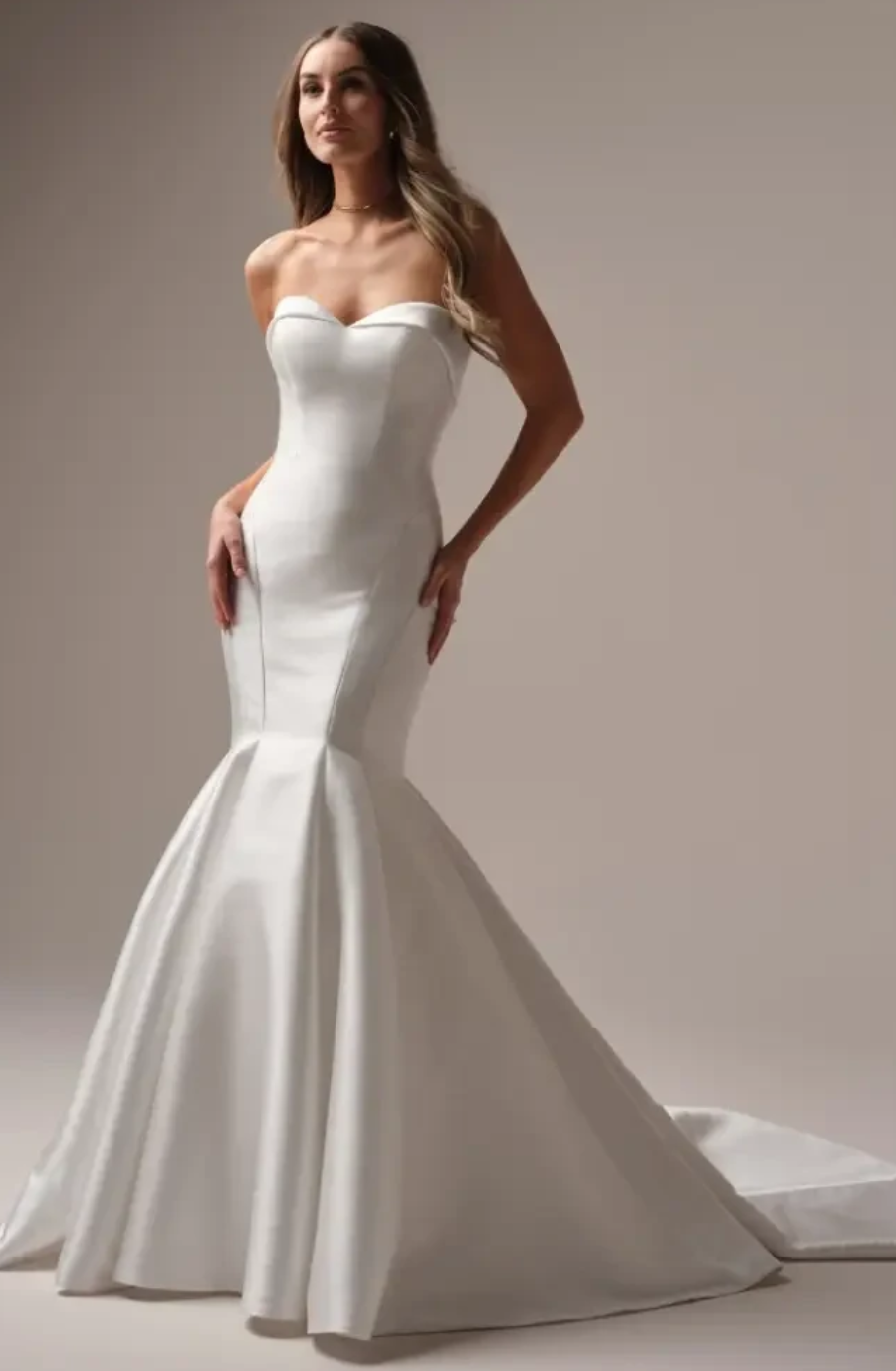 A model is wearing a fitted strapless white wedding gown with a mermaid silhouette and a long train.