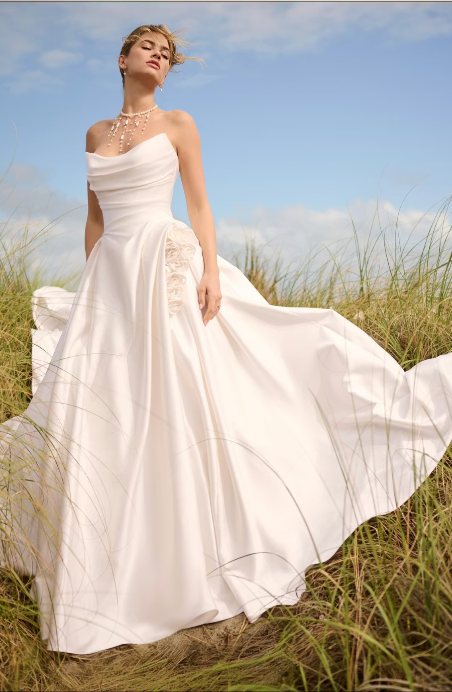 A model wearing a flowing white wedding dress stands gracefully in a field with tall grass and a blue sky.