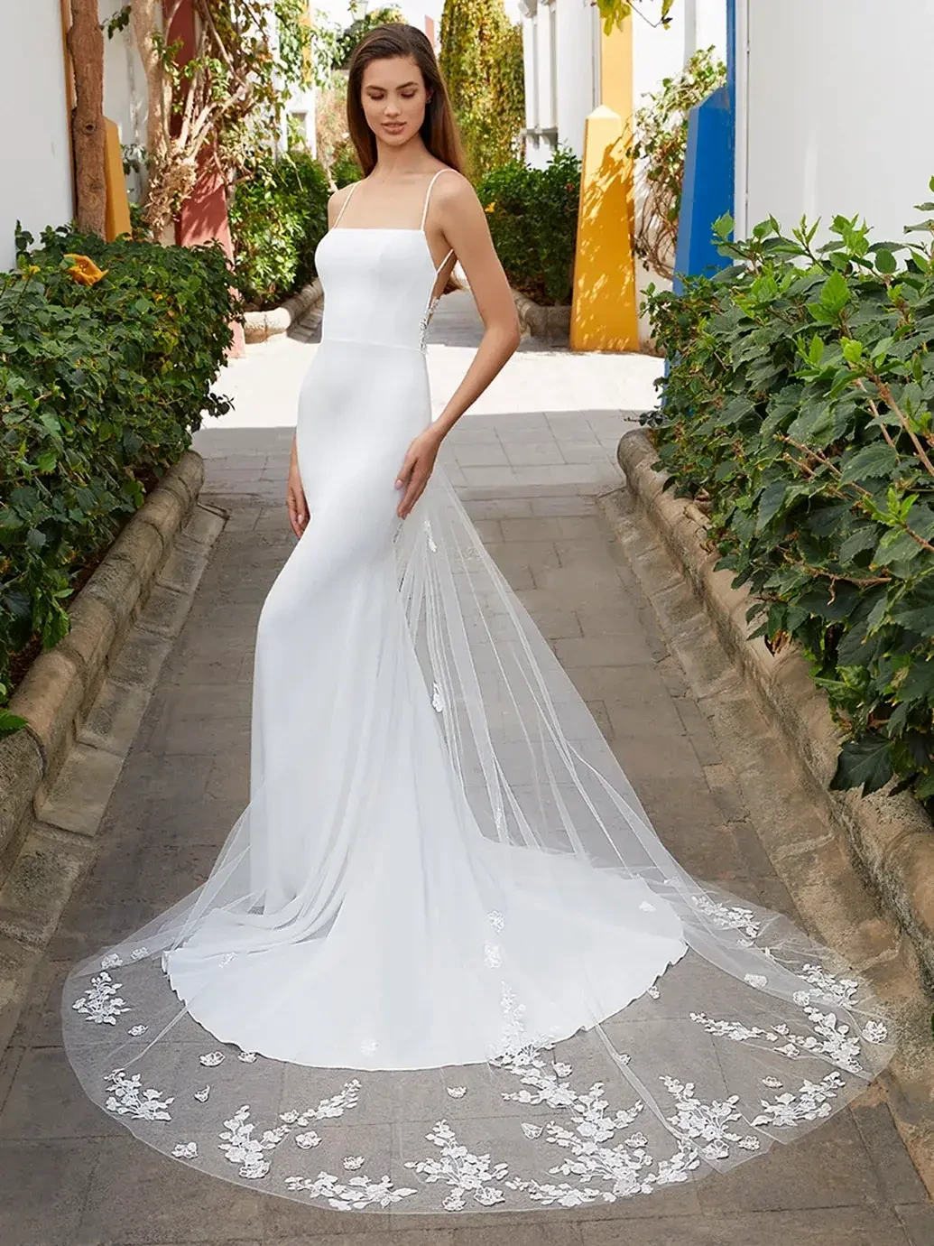 Model is wearing a lightweight stretch georgette gown with thin spaghetti straps, a modern square neckline, and a low illusion back detailed in delicate floral lace.