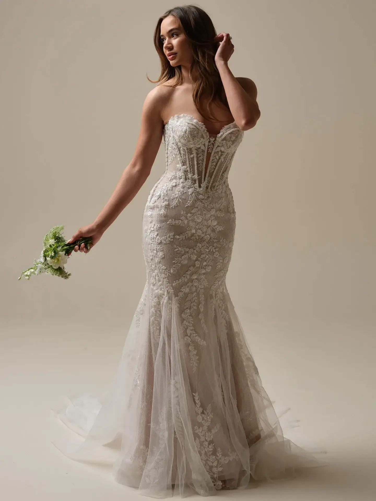 A woman in a strapless, lace-embroidered wedding gown with a fitted silhouette holds a small bouquet. She looks to the side with a serene expression.