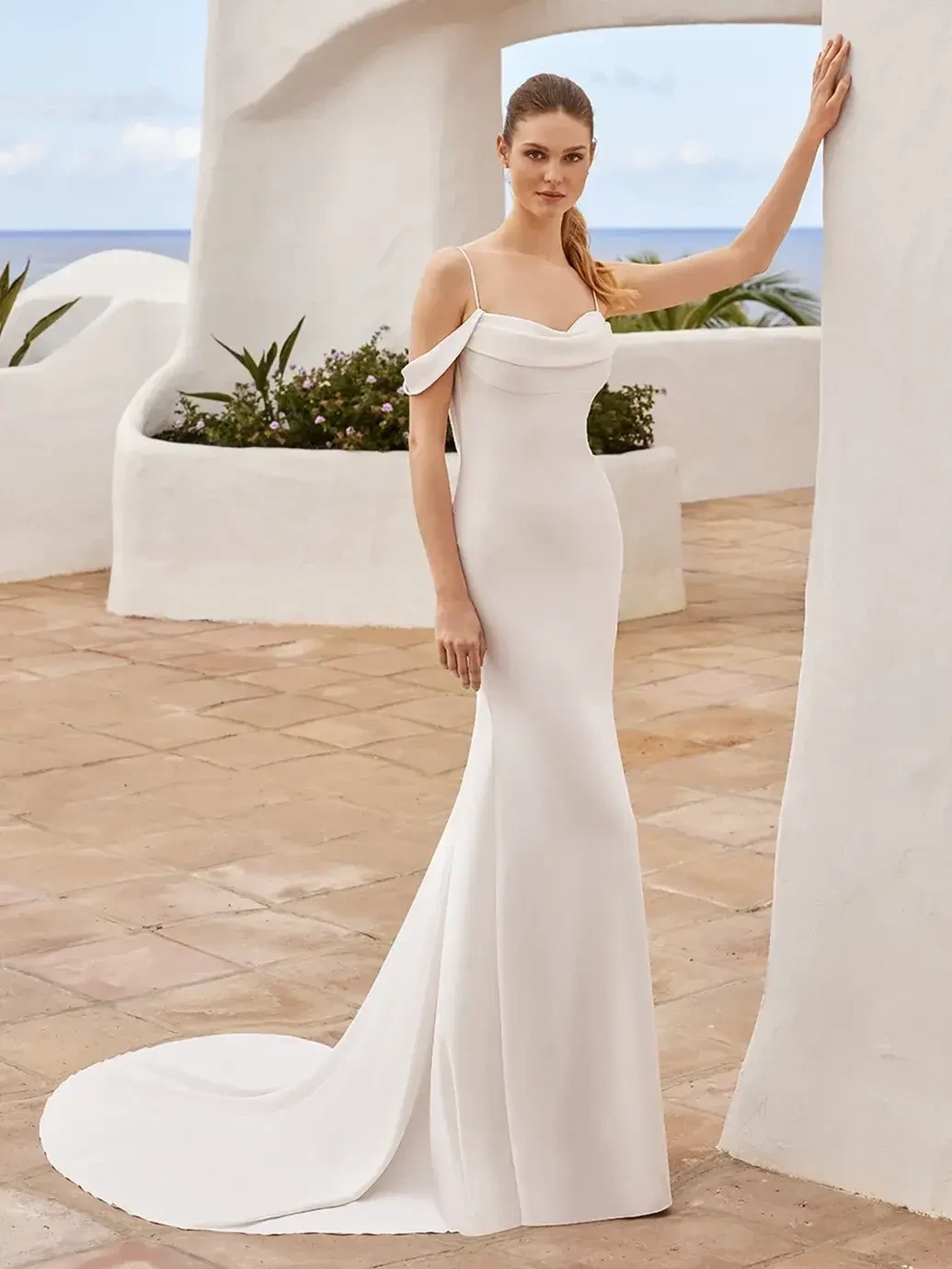 Model is wearing a sleek crepe gown with delicate spaghetti straps, a low scoop back, and a flowing sash