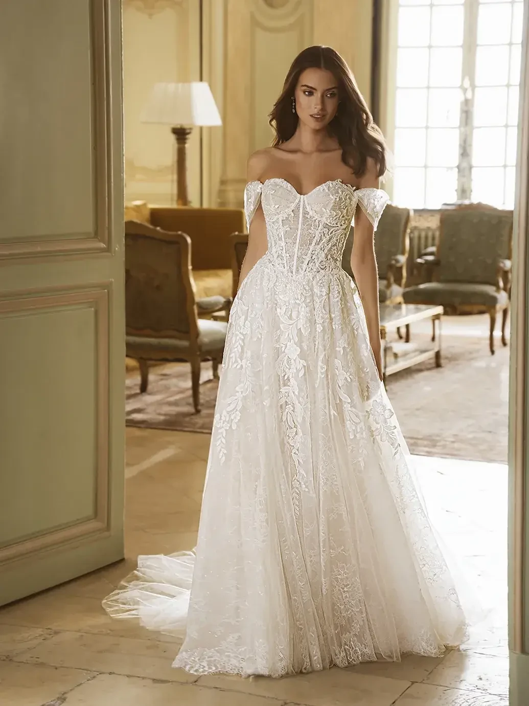 A woman is wearing an off-the-shoulder white wedding dress with intricate lace detailing, standing in a beautifully decorated room with elegant furnishings.