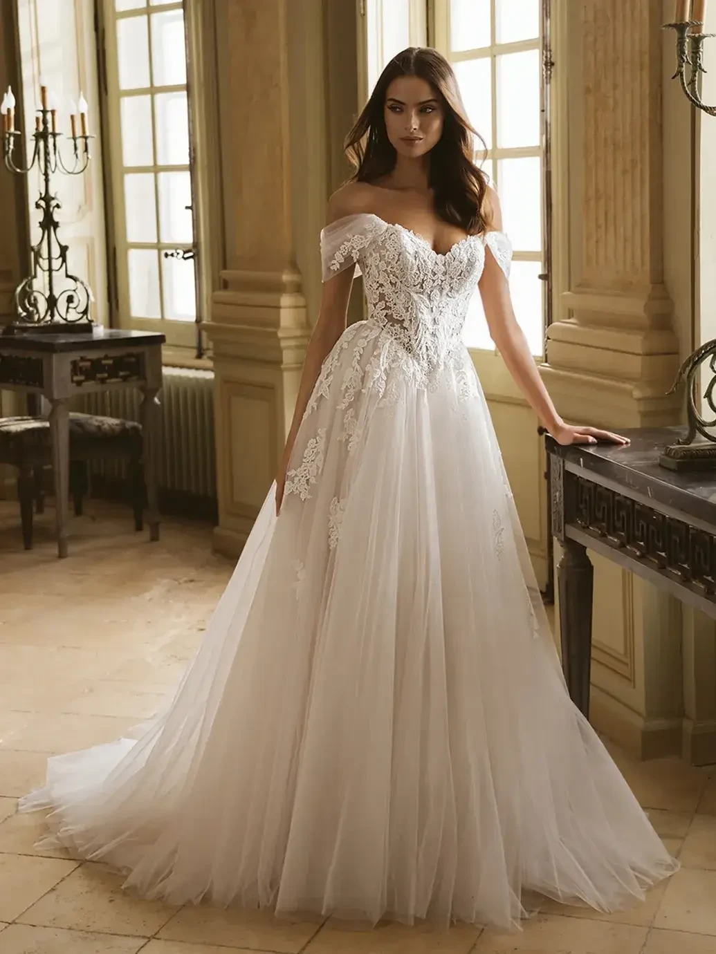 A model is wearing a beautiful off-the-shoulder wedding dress with a fitted lace bodice and a flowing tulle skirt, standing in an elegant room with large windows and soft lighting.