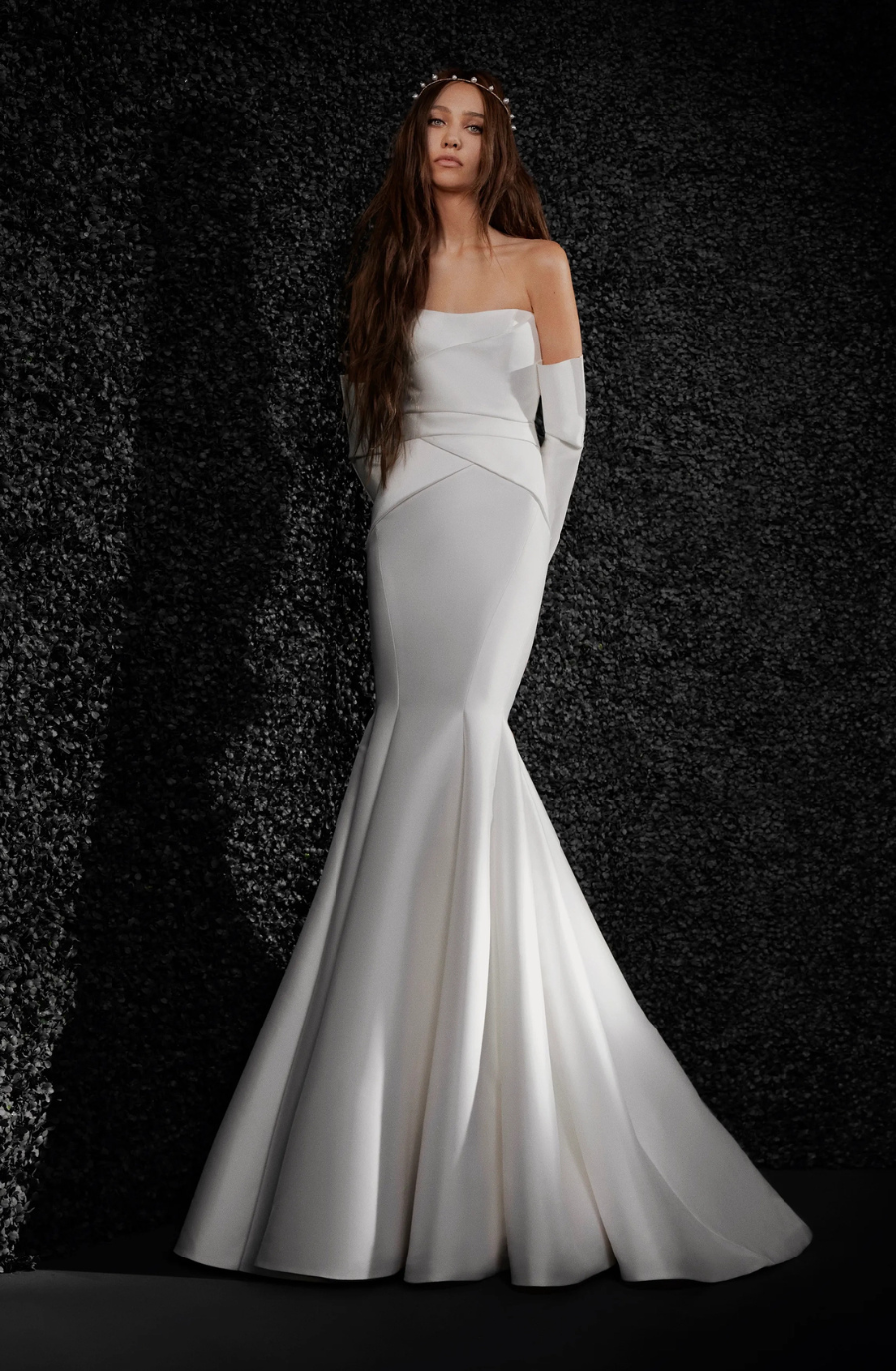 A woman in a strapless, form-fitting white gown with long gloves stands against a dark, textured background. Her long hair flows naturally, conveying elegance.