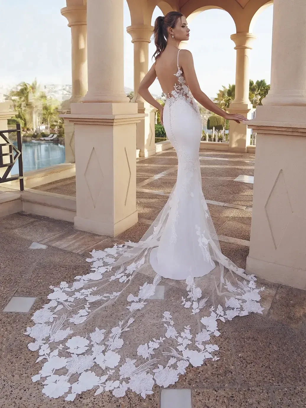 Model is wearing a form-fitting gown with a lace bodice, sleek skirt, open illusion back, and dramatic lace-detailed train