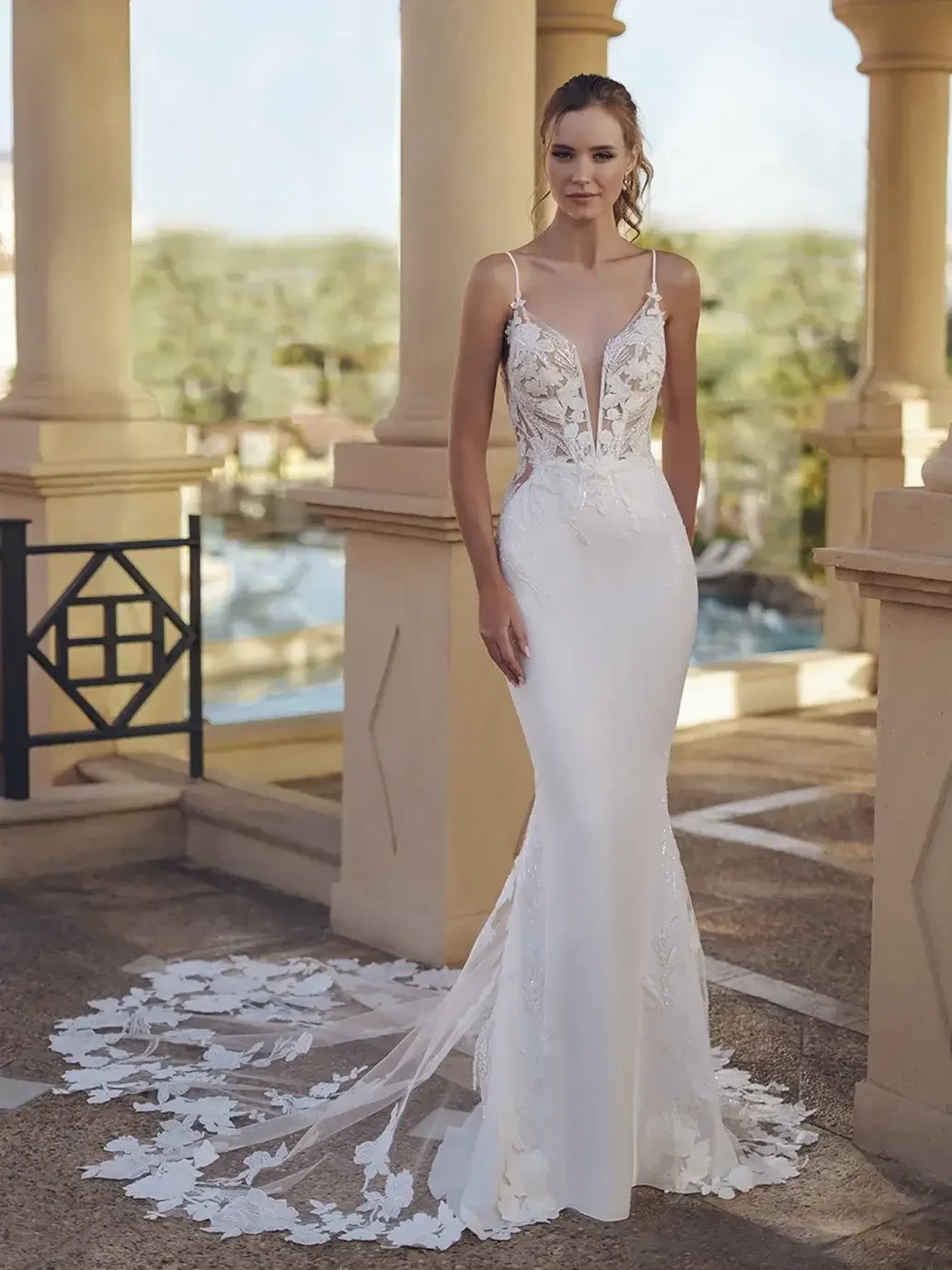 Model is wearing a form-fitting gown with a lace bodice, sleek skirt, open illusion back, and dramatic lace-detailed train