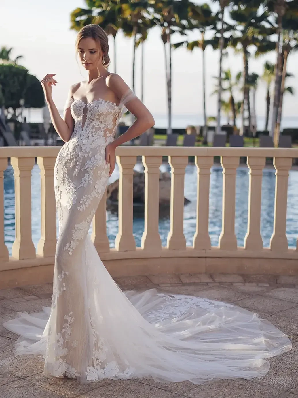 Model is wearing a romantic off-the-shoulder gown with 3D floral appliqués, a sculpted lace silhouette, and a sheer train