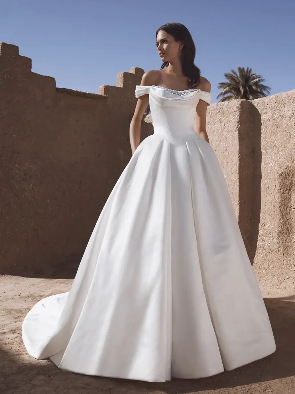 A woman wearing an off-the-shoulder white gown stands against a textured, earthen wall with palm trees in the background.
