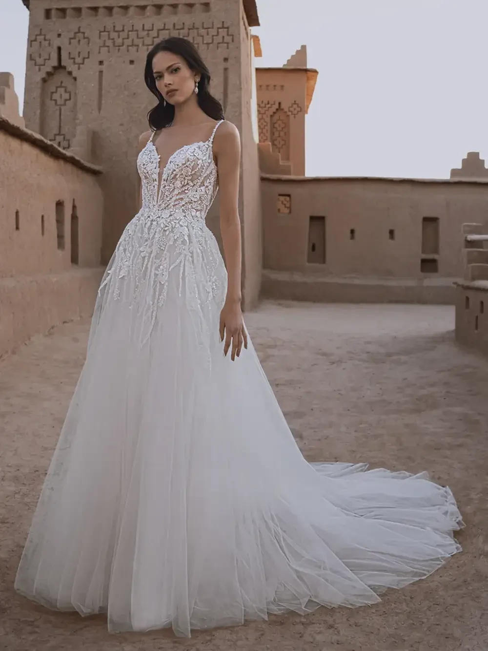 A woman in a beautiful, intricate white wedding dress standing in a sandy courtyard with traditional architecture in the background.