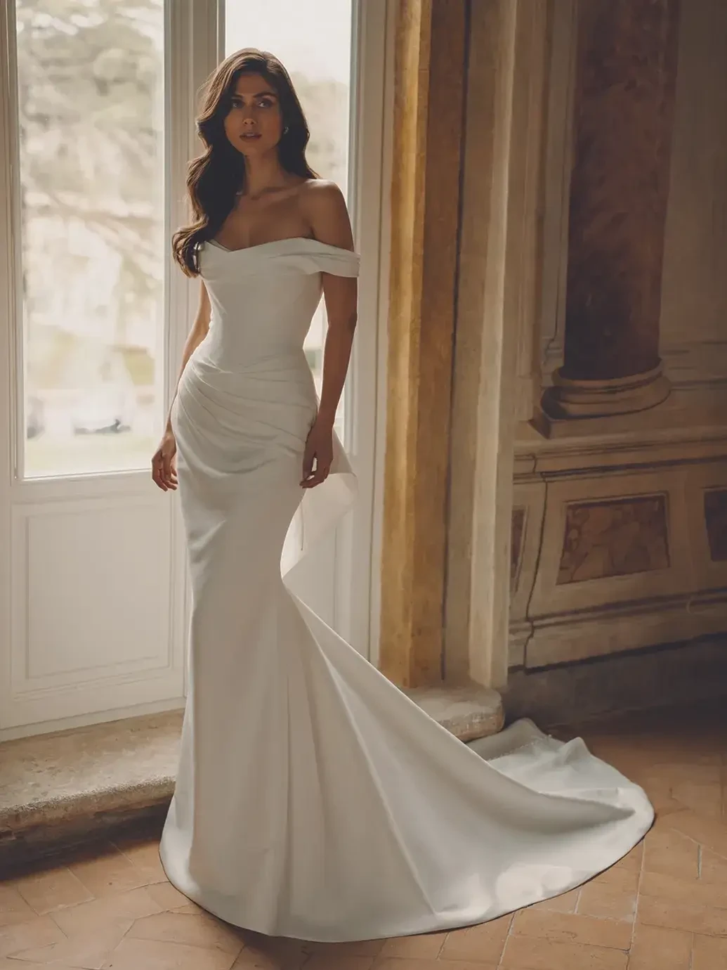 A woman standing in a bright room wearing a sleek, off-the-shoulder white gown with a fitted silhouette and a dramatic train. She has long hair and is gazing elegantly out of a window.