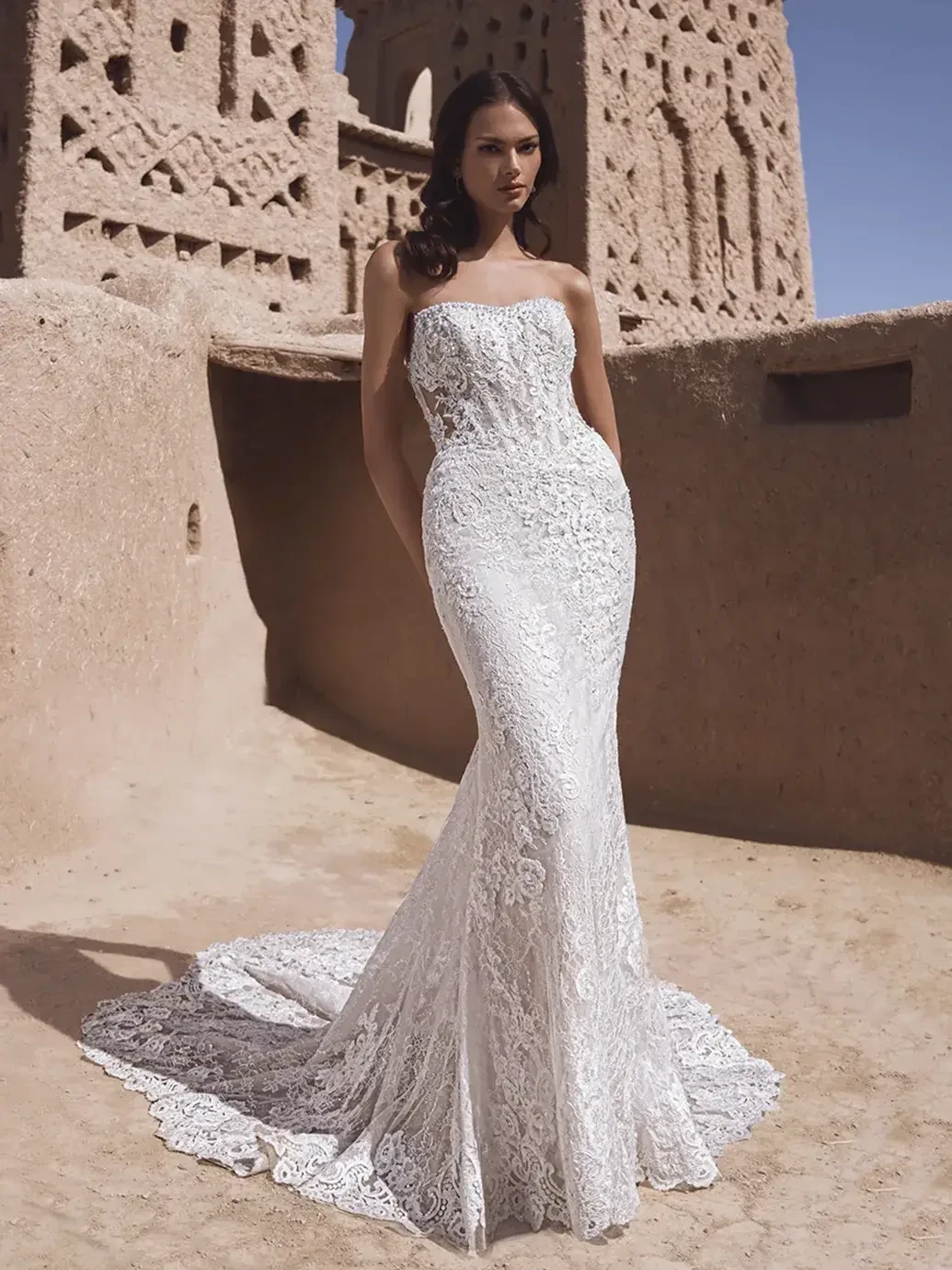 A model wearing a form-fitting white lace bridal gown stands in an outdoor setting with ancient architecture in the background. The dress features a strapless design and a flowing train, accentuating the model's silhouette.