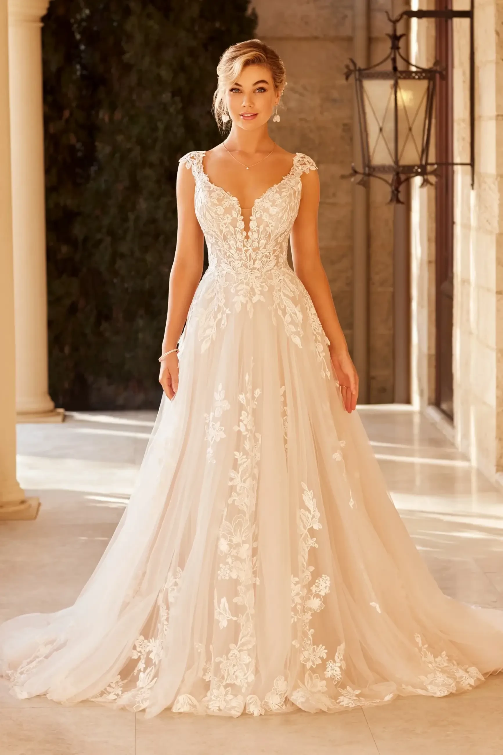 A woman in a flowing, lace-embroidered wedding gown with a deep V-neck stands in a sunlit, elegant hallway. The scene exudes a romantic and joyful tone.