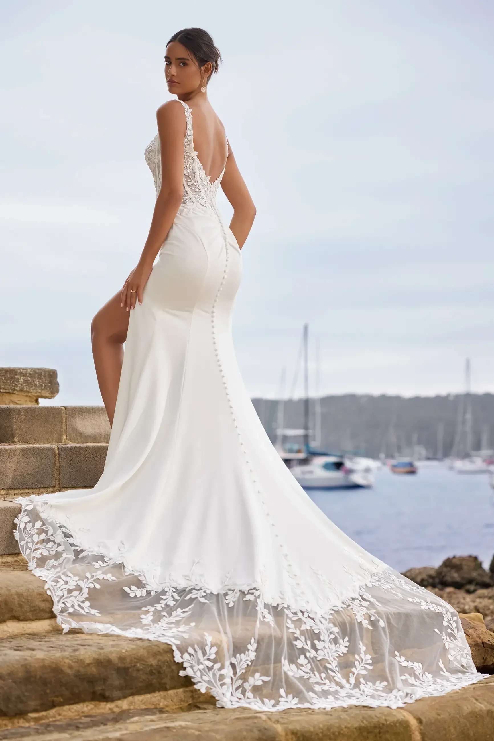 Model is wearing a fit-and-flare satin crepe wedding dress with a thigh-high slit, floral lace corset bodice, lace straps, and a deep V-back with fabric-covered buttons and a lace train.