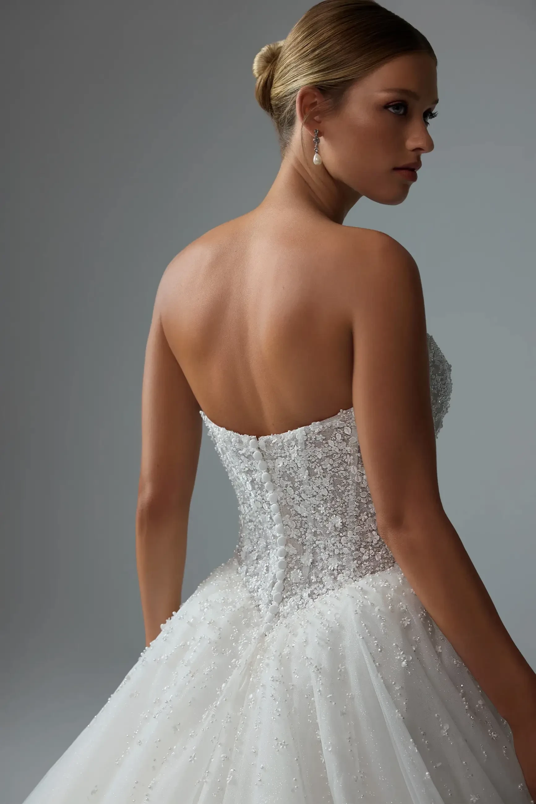 Model is wearing a strapless ball gown with a semi-sheer V-neck bodice adorned with pearls and 3D floral lace, a basque waist, and fabric-covered buttons down the back.