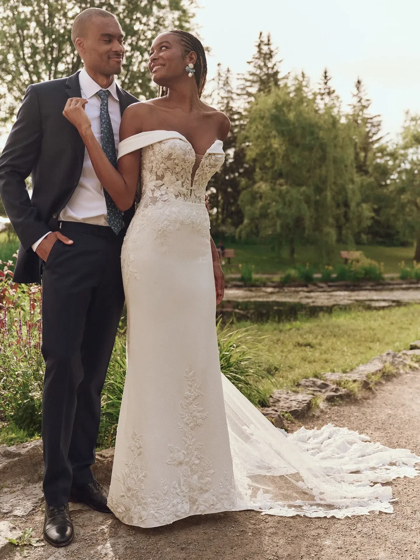 A groom in a dark suit and bride in an off-shoulder lace gown smile at each other in a lush garden setting, exuding joy and love.