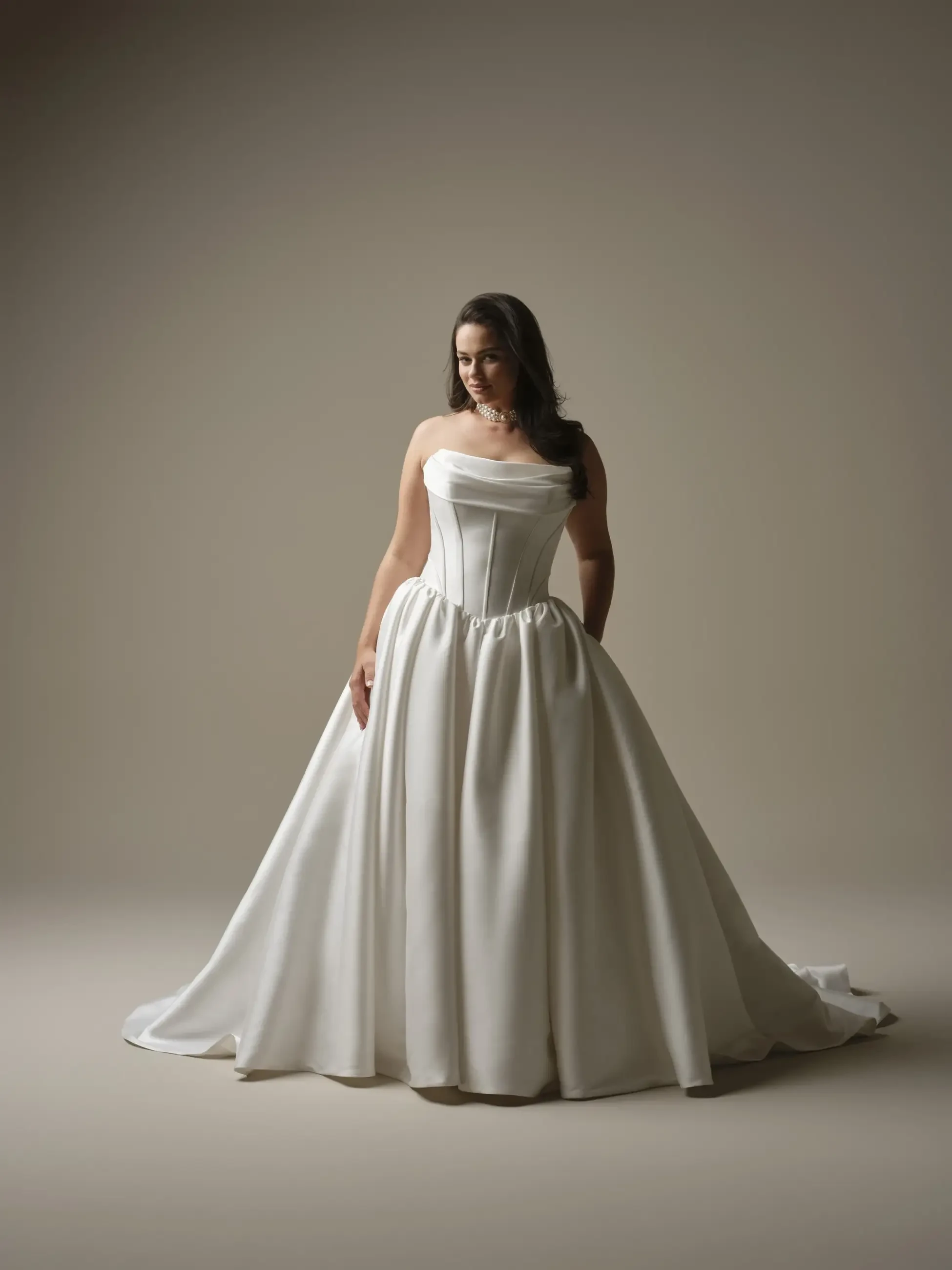 A woman stands in a strapless, flowing white gown, exuding elegance. She gazes downward, creating a serene, graceful atmosphere in a softly lit studio.
